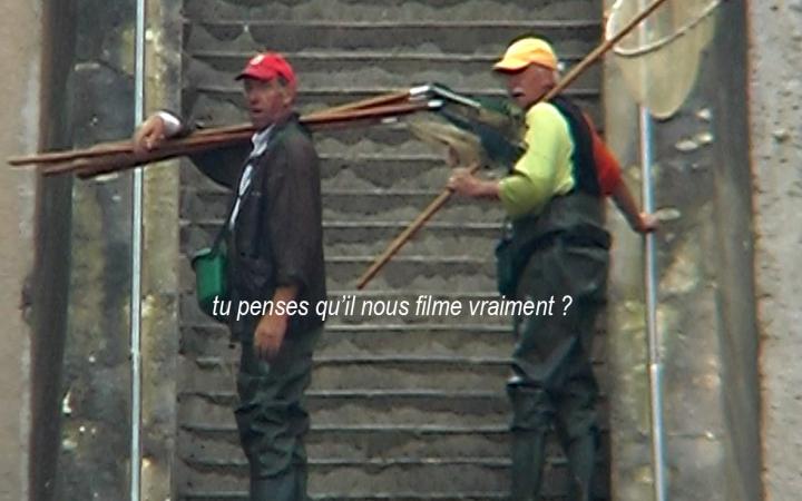 A film still from the film Héros absurde by Edmund Kuppel shows two men at the foot of an outdoor stone staircase, one on the left, the other on the right. They wear caps and fishing gear. They ask: "Tu penses qu'il nous filment vraiment?"