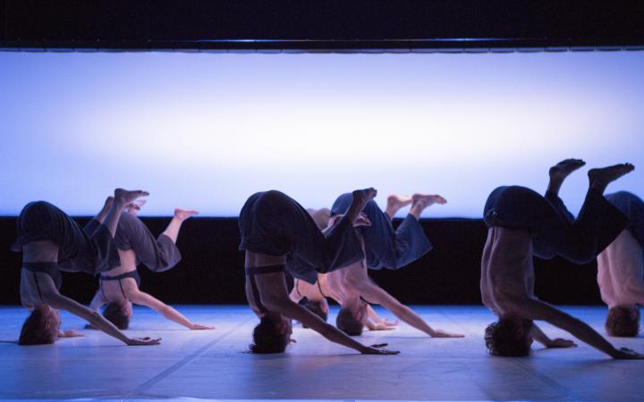 Seven different dancers have bent their legs in the headstand. In the background there is a large, milky screen. 