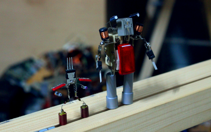 Auf einem Holzbalken stehen zwei aus Elektronikschrott zusammengelötete Roboter.