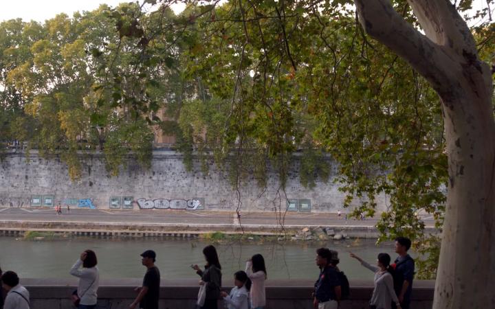Film still by Marijke van Warmerdam. You see a riverbank in Rome. On the right a tree and on the left people walking along the riverbank.