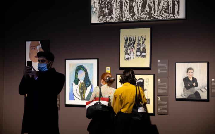 Zu sehen sind Besucher:innen, die sich von Überlebenden gemalte Kunstwerke sowie Porträtfotos anschauen