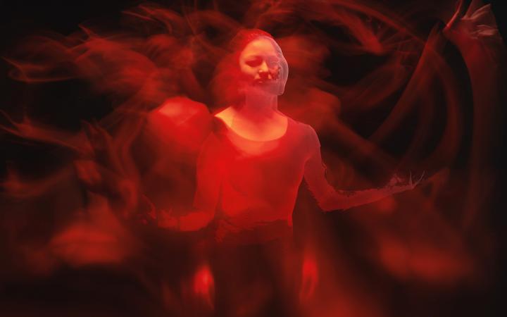 The photo shows a woman illuminated by red light in a darkened room. This expression shows several blurred arms and hands in motion, almost resembling an Indian deity. 