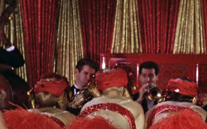 Zu sehen sind zwei Trompetenspieler vor einem goldenen und roten Vorhang. Im Publikum sitzen drei Frauen mit rotem Kopfschmuck