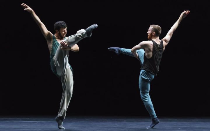 Two dancers of the choreography »Friends of Forsythe«. They stand opposite each other on a black stage and artfully raise their arms and legs in the air.