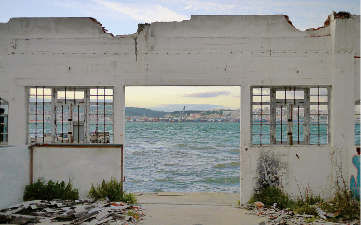 Das Bild zeigt eine Hausruine, durch deren offene Türen und Fenster man auf das Meer und eine dahinter liegende Stadt sehen kann. 