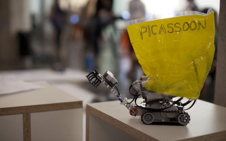 Auf einem Tisch steht ein kleiner Lego-Roboter, an dem ein gelbes Schild mit dem Wort "Picassoon" befestigt ist.