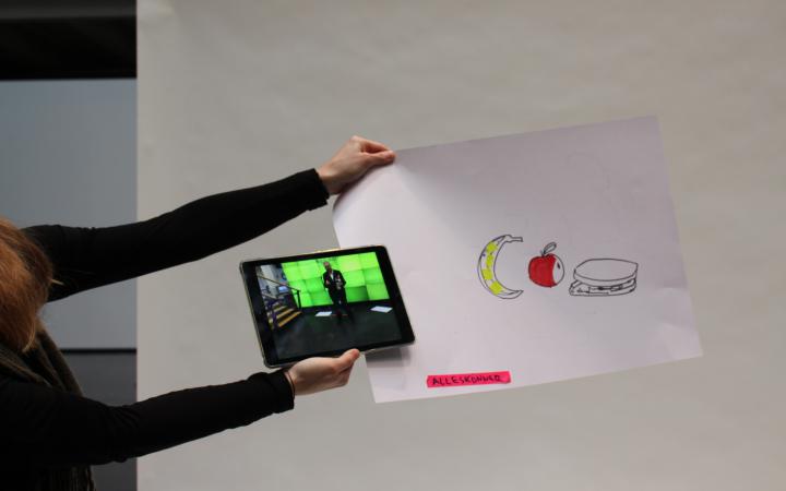 A woman is holding a piece of paper on which a banana, an apple and a sandwich are sketched. On the bottom of the paper the word "Allrounder" written. Also she is holding an iPad with the photo of a man in front of a green screen.