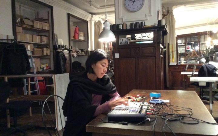 Eine junge Frau sitzt an einem Tisch vor verschiedenen technischen Geräten. Sie kreiert elektronische Musik.