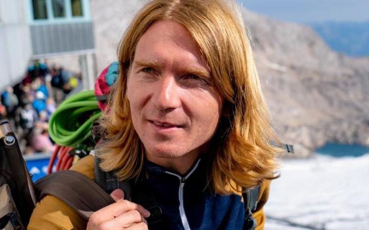 The picture shows the artist Markus jeschanuig. He is pictured outside and seems to be on a hike on a mountain top. He wears open shoulder-length blond hair and looks left out of the picture.