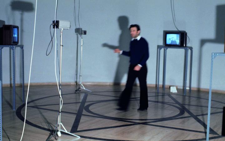 A man stands in a pentagram surrounded by monitors and cameras. The photo shows Peter Weibel as part of the exhibition »respektive Peter Weibel«.