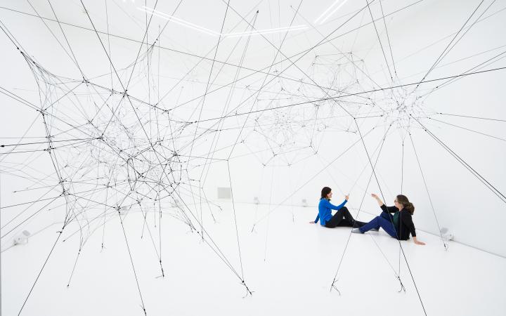 »Algo-r(h)i(y)thms« von Tomás Saraceno. Zu sehen ist ein großes Netz aus Fäden in einem weißen Raum. Zwei Personen sitzen in diesem Raum und berühren diese Fäden.