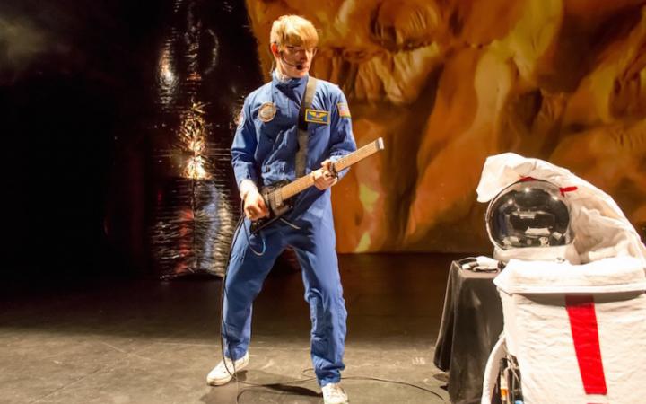 Zu sehen ist ein junger Mann in einem blauem Astronauten Anzug und einer Gitarre in der Hand.
