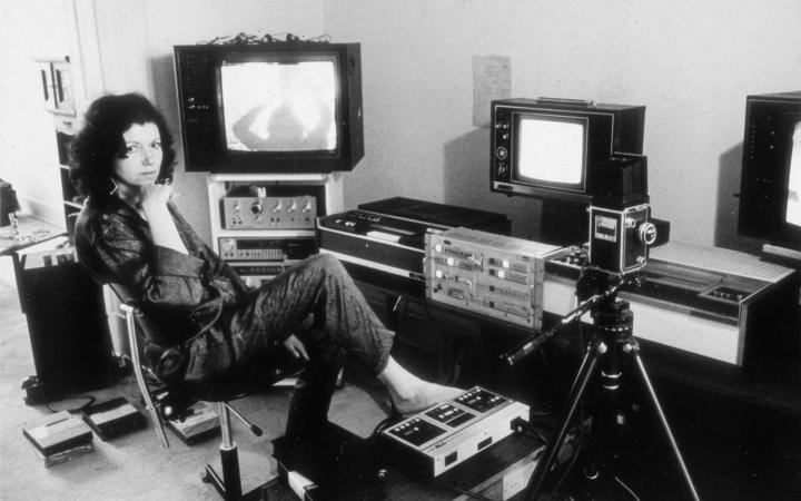 Das Foto ist schwarz-weiß und zeigt Ulrike Rosenbach, die Medienkünstlerin, in ihrem Studio in den 70er Jahren. Ulrike Rosenbach sitzt auf einem Drehstuhl und ist umgeben von Kameraequipment und Röhrenbildschirmen. 
