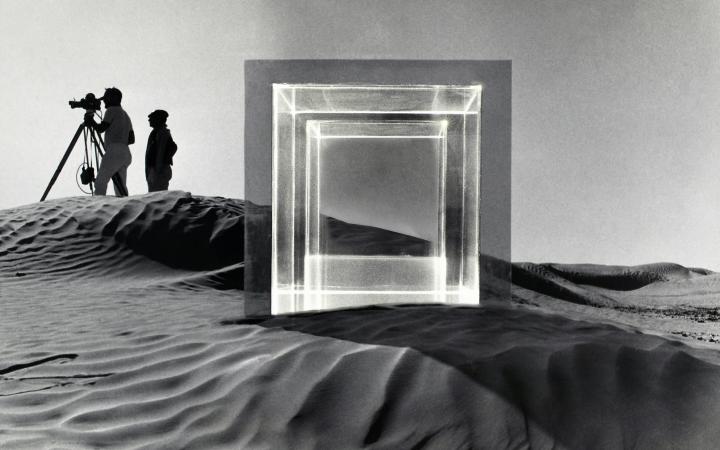 In a desert landscape around 1968, filming of the movie "Tele-Mack" took place. In the foreground is a large square glass sculpture and in the background are two men with a film camera. The picture is black and white.