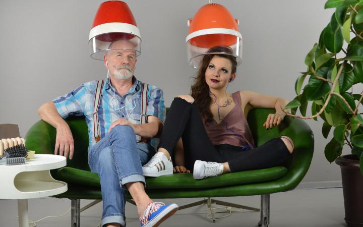 Two people sit on a green sofa, their heads are stuck under a hairdresser's hood.