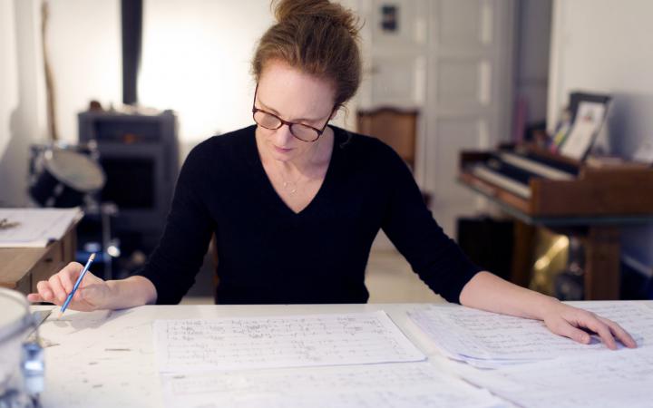 Photo of a woman with glasses and reddish hair, studying a sheet of music lying on a table.