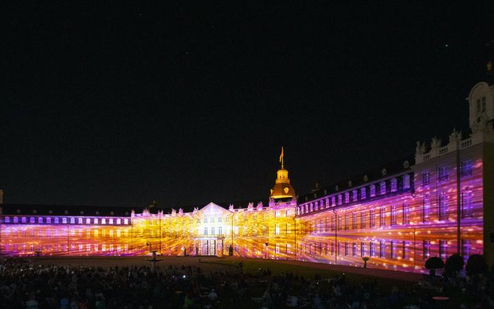 Die Fassade des Karlsruher Schlosses ist in bunten Farben angestrahlt. Es ist Nacht.