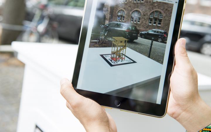 Auf einem Tablet-Bildschirm ist eine virtuelle Skulptur zu sehen, den realen Raum eingebettet ist.