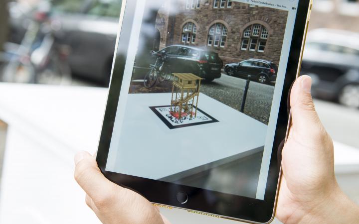 Two hands hold an iPad on which a 3D animated miniature monument can be seen over the QR code of the work »The Peoples Monuments«..