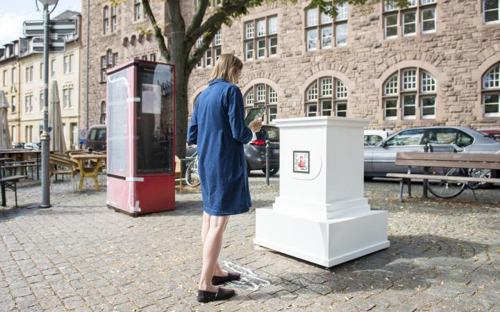 A woman in a blue dress stands in front of an empty white pedestal. The pedestal belongs to the AR artwork »The People's Monuments«.