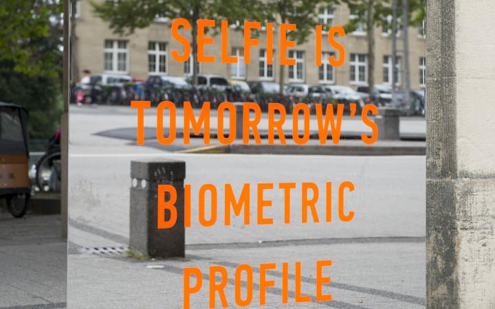 Zu sehen ist ein orangener Schriftzug »Todays Selfie is Tomorrows Biometric Profile« ist auf einem großen rechteckigen, rahmenlosen Spiegel. Die Installation steht draußen vor dem Eingang-Süd des Zoos.
