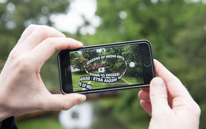 Zwei Hände halten ein Iphone auf dem ein App-Kunstwerk geöffnet ist. Nun sieht man die kleine Brücke im Zoo über der die Ausstellungs-Schlaufe schwebt.