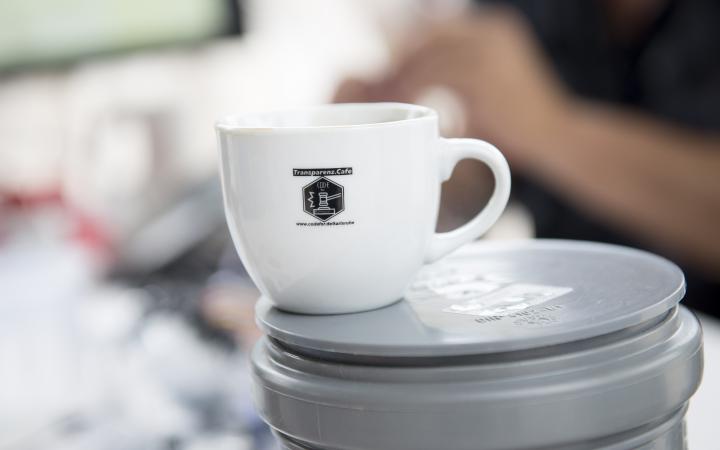 You can see a white coffee cup with the Transparenz Cafè logo on it.