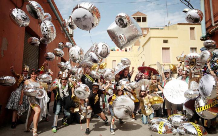 A group of men and women is standing on the street and each one is holding a silver balloon. On the balloons are pictures of faces and eyes. The people are cheering and smiling into the camera.