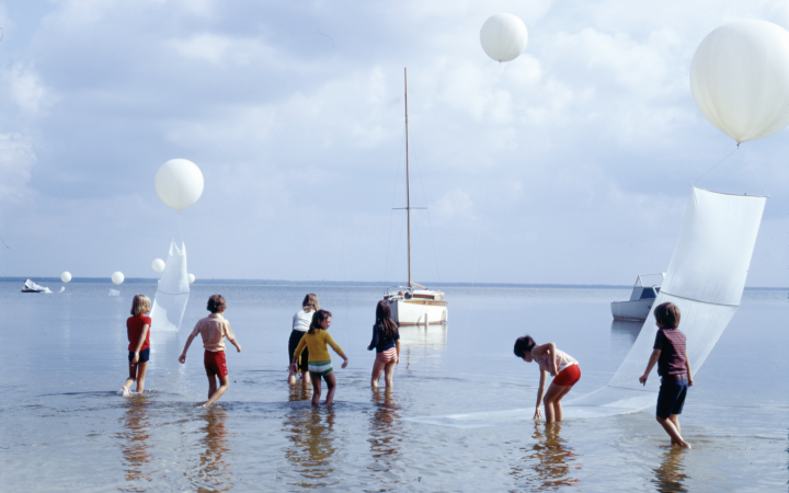 You can see several children running through water. Above them are white balloons. In the background you can see boats. 