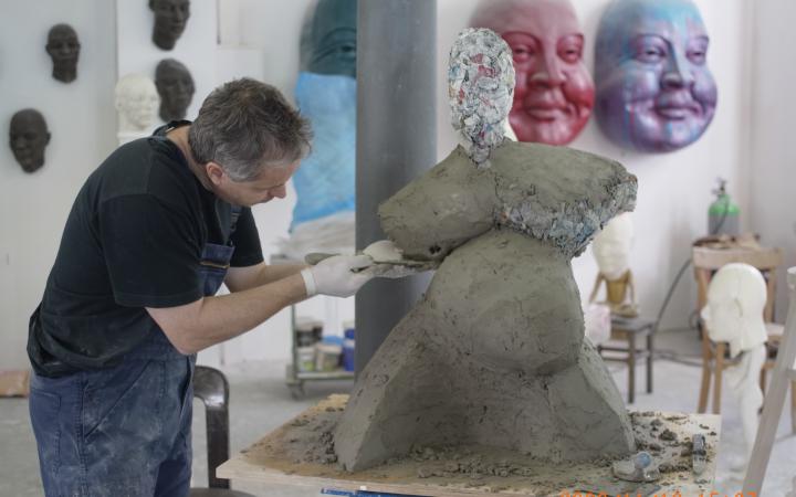 Ein Mann in Latzhosen modelliert eine voluminöse Figur aus Ton.