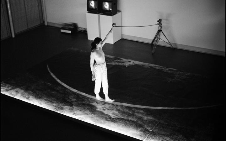 Ulrike Rosenbach, »Die einsame Spaziergängerin«, 1979. Zu sehen ist eine Person auf einer Bühne, die barfüßig auf einer gebogenen Linie läuft. Dabei hält sie ein Seil in der Hand, das an einem Stativ befestigt ist. Das Bild ist schwarz-weiß. 