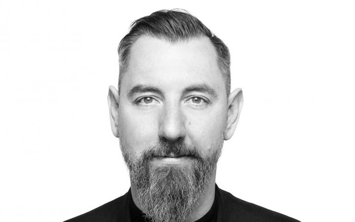 You can see a portrait of Stephan Schwingeler. it is a black and white photo and shows him from the chest up. He wears a beard and a black T-shirt and looks frontally into the camera.