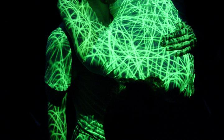 Kenneth Flak stands upright, in his left arm he holds Külli Roosna, she hugs him. Both are covered by a projection with neon green lines.