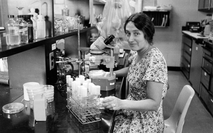 The young Lynn Margulis is sitting in a laboratory with numerous laboratory utensils. She is wearing a summer dress and smiles slightly.