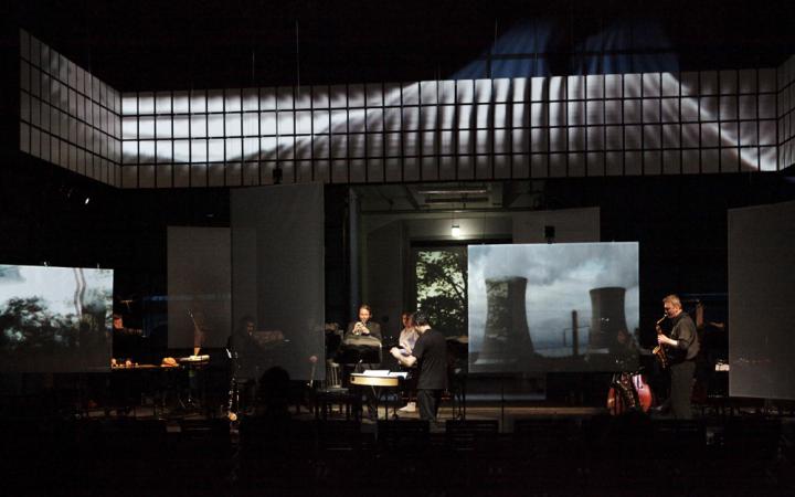  Ein Musikensemble auf einer dunklen Bühne. Im Hintergrund ein Bild von einem Atomkraftwerk