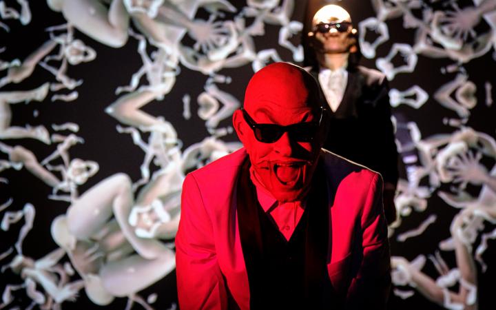 Im Vordergrund ist ein rot beleuchteter Mann zu sehen. Die obere Hälfte seines Gesichts ist mit einer Maske bedeckt in der Form eines Gesichts. Er trägt eine Sonnenbrille und einen Anzug. Im Hintergrund sind abstrakte Formen erkennbar.