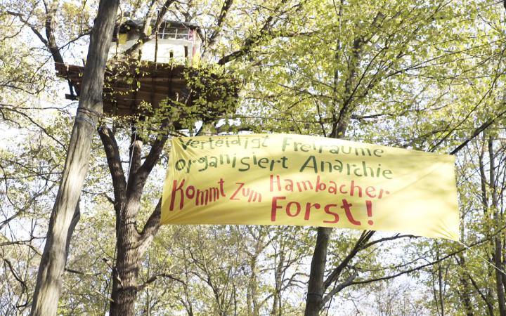 Zu sehen ist ein Baumhaus. Unterhalb des Baumhauses ist ein Banner zwischen den Bäumen gespannt auf dem zu lesen ist: »Verteidigt Freiräume. Organisiert Anarchie. Kommt zum Hambacher Forst!«