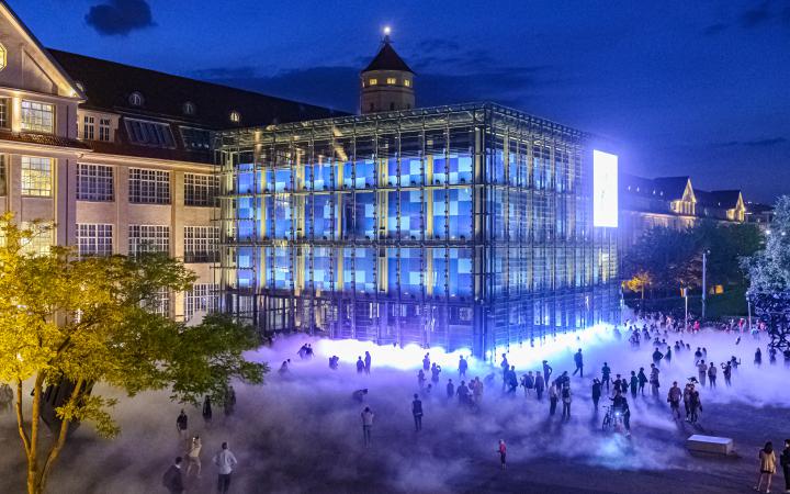 The photo was taken at night with a drone. It shows the fog sculpture and many silhouettes of visitors of KAMUNA 2019. The cube and the facade of the building are brightly illuminated.  