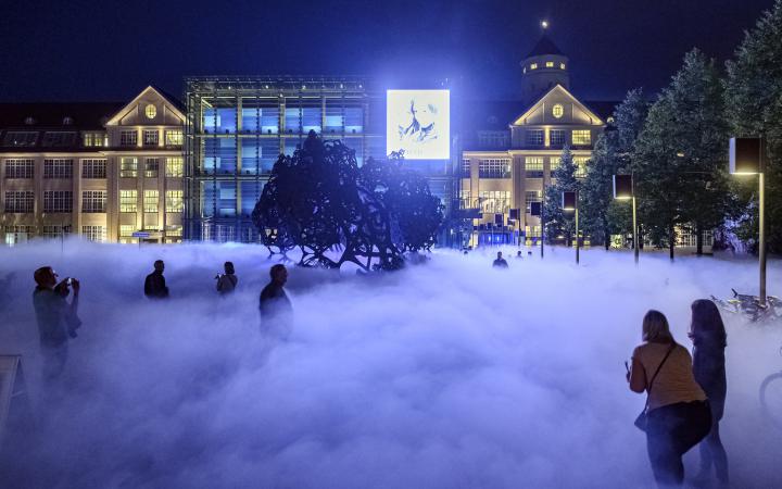 Auf dem Foto sieht man die Nebelskulptur bei Nacht. Das Foto zeigt den die hell erleuchtete Fassade des ZKM Gebäudes und man sieht den Mittelteil mit Kubus und Osteingang 