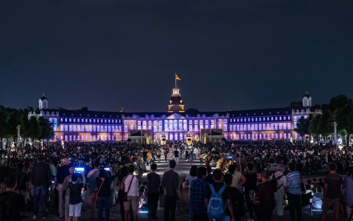 The black sky as a background highlights the brightly lit castle. Karlsruhe Castle illuminates in various shades of purple and pink, exploiting the structures of the castle to create geometric patterns.