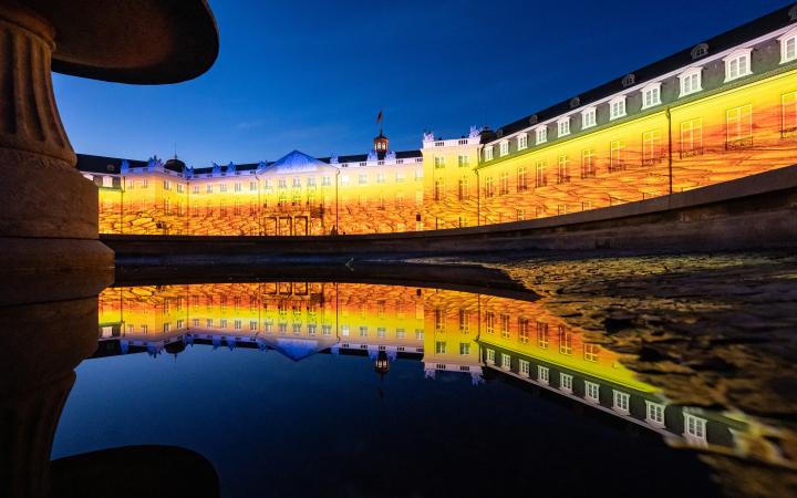 The castle of Karlsruhe illuminates in yellow-orange hues. In the foreground the facade is reflected in the water of the fountain. The evening sky has already darkened.