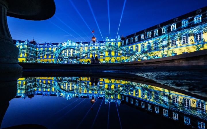 Zu sehen ist die beleuchtete Fassade des Karlsruher Barockschlosses in den Farben Blau und Gelb.
