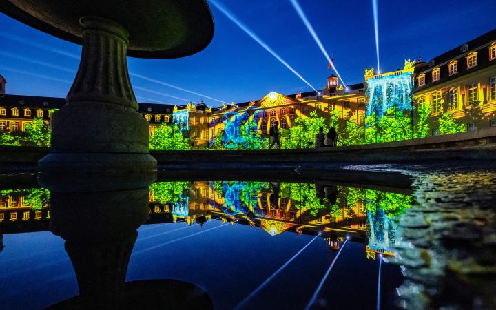 In the background you can see the illuminated facade of Karlsruhe Castle. A blue sky is projected over a landscape of trees with a waterfall. In the foreground the projection is reflected in a fountain. Spotlights rise into the sky.