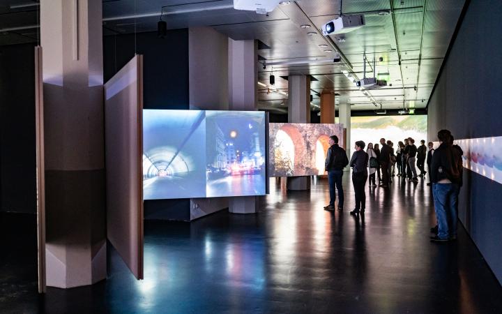 Exhibition view »Marijke van Warmerdam. Then, now, and then«. A darkened room can be seen. Several screens hang from the ceiling and show different recordings. People are looking at these recordings.