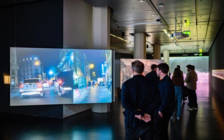 Zu sehen sind zwei Personen vor einem Bildschirm, auf dem ein Auto auf der Straße gezeigt wird.Ausstellungsansicht »Marijke van Warmerdam. Then, now, and then«.