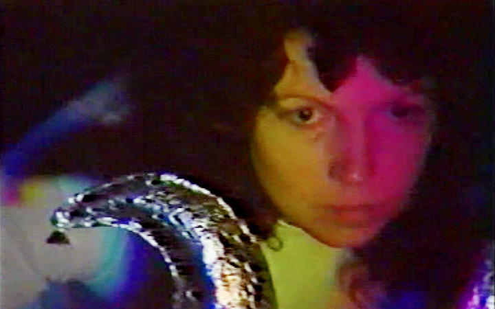 Man sieht Ulrike Rosenbach, die Medien-Künstlerin, in ihrem Performance Werk die Eulenspieglerin im Jahr 1985. Das Videostill zeigt das Gesicht von Ulrike Rosenbach mit schwarzen Locken und hinter einem bunten Filter. 