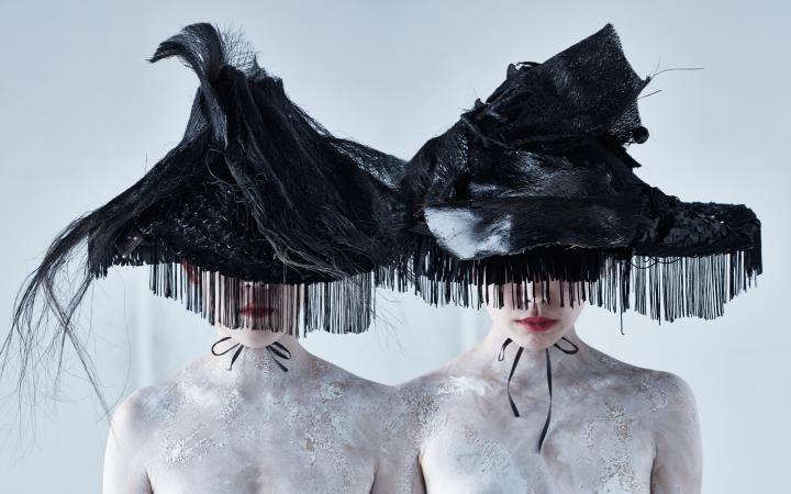 Porträt von zwei jungen Frauen, deren Augenpartien durch übergroße, dreieckige, schwarze Hüte verdeckt werden 