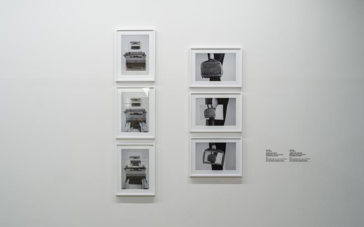 On view are six picture-in-picture, black and white photographs in picture frames on a wall. Three of them show a typewriter, the other three show a hand holding a briefcase.