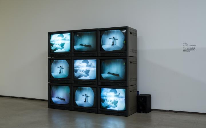 Zu sehen sind neun kleine Röhrenfernseher, angeordnet als Quadrat. Sie zeigen eine nackte Person in verschiedenen Positionen in einem leeren Raum.