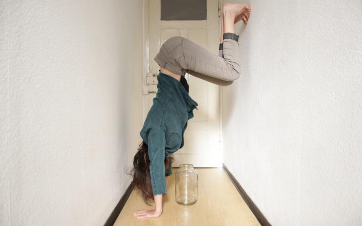 A person is doing a handstand against the wall in a small hallway, under her is an empty jar.